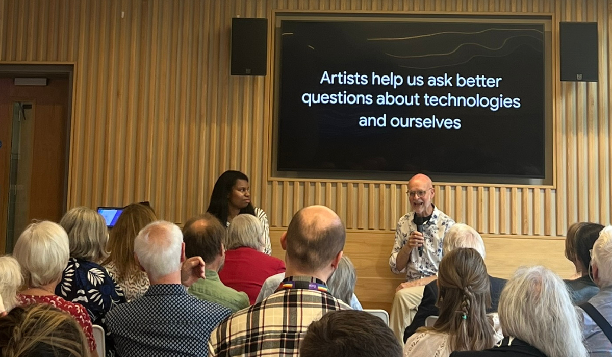 Panel event with Pamela and Alan in the cafe, in front if seated audience. The screen behind them displays a message that reads: "Artists help us ask better questions about technologies and ourselves." 