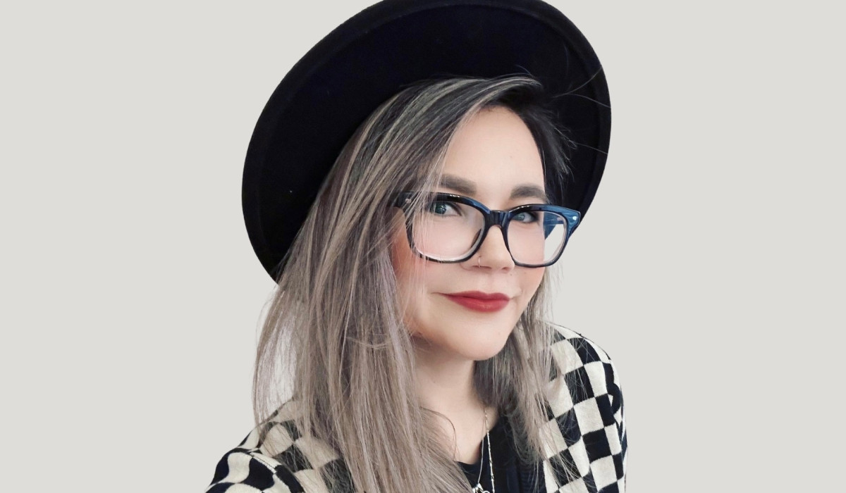 Amy De la Force headshot. Amy is wearing a black hat and a checkered jumper