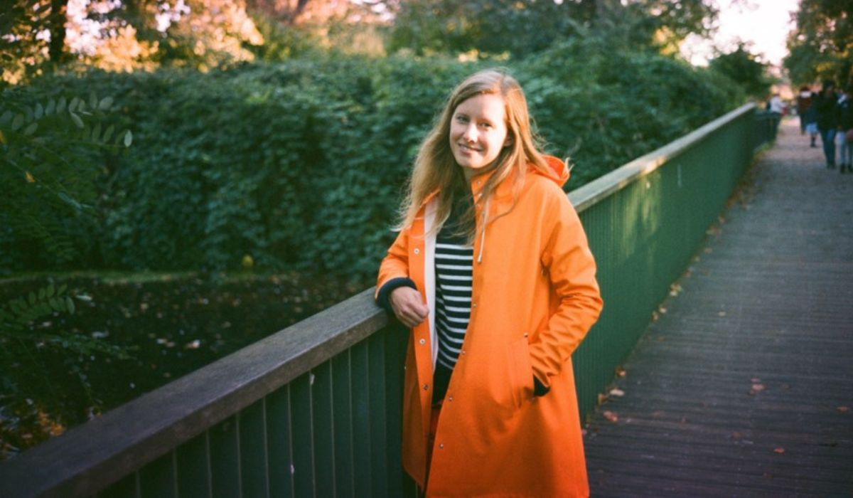 Matilda posing for picture on a bridge surrounded by greenery. She is wearing an orange raincoat and stripey tshirt