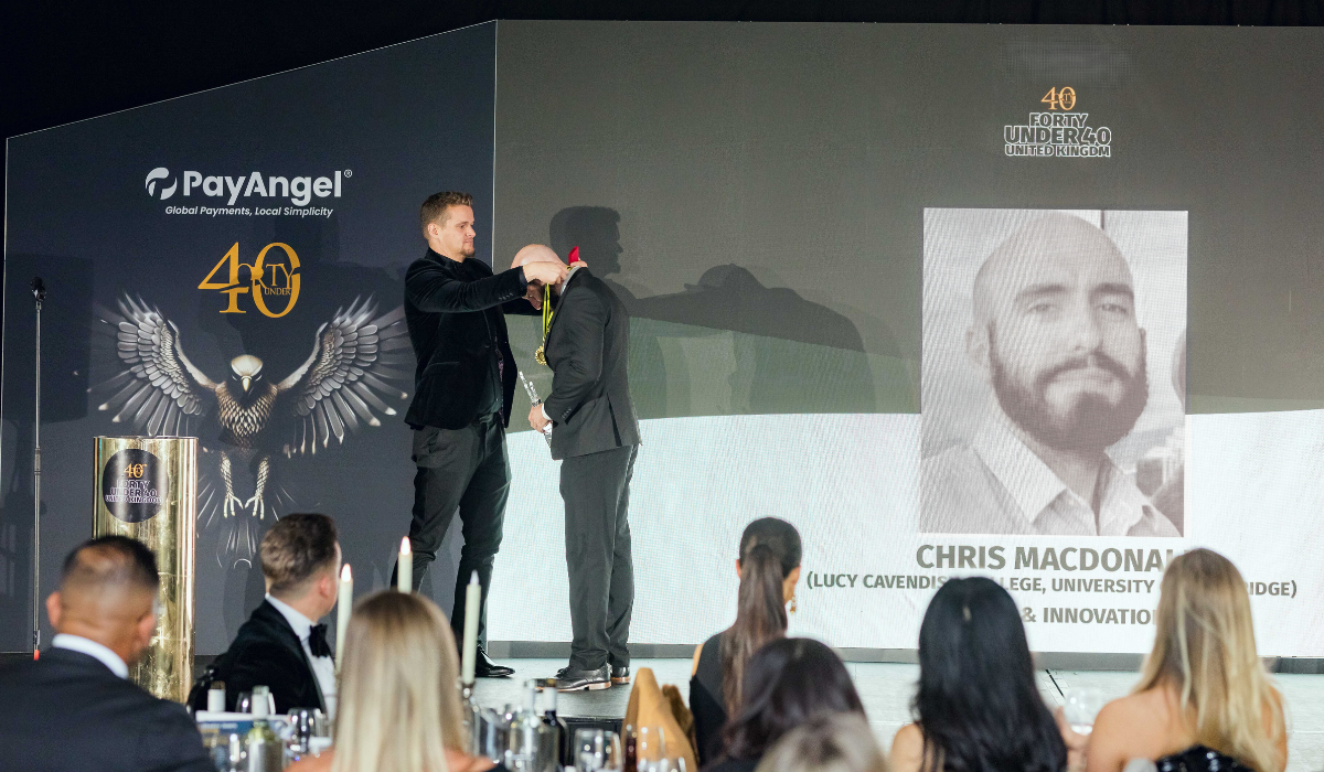 Chris is receiving a medal on stage at the awards ceremony. Behind them is a large screen displaying the name CHRIS MACDONALD and his black-and-white portrait. The text on the screen also mentions "LUCY CAVENDISH COLLEGE, UNIVERSITY OF CAMBRIDGE" and "& INNOVATION". The backdrop includes the logo for PayAngel and the event branding 40 UNDER 40 UNITED KINGDOM with an eagle motif. In the foreground, there are seated guests dressed formally, watching the stage.