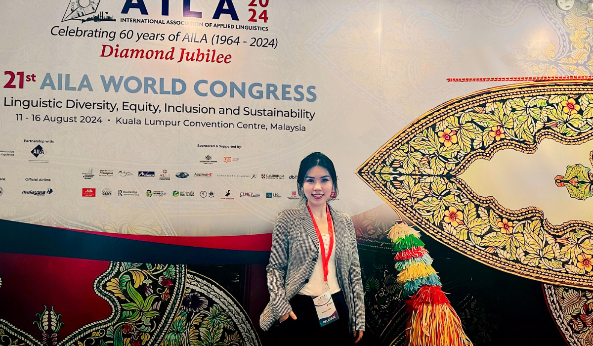 Mengxi standing in front of a large conference backdrop that reads "21st AILA World Congress: Linguistic Diversity, Equity, Inclusion, and Sustainability," held at the Kuala Lumpur Convention Centre, Malaysia, from August 11 to 16, 2024. The event celebrates 60 years of AILA (1964–2024) as part of its Diamond Jubilee. The backdrop features vibrant traditional Malaysian artwork and logos of sponsors and partners. The woman is dressed professionally in a plaid blazer and white blouse, wearing a red lanyard wi