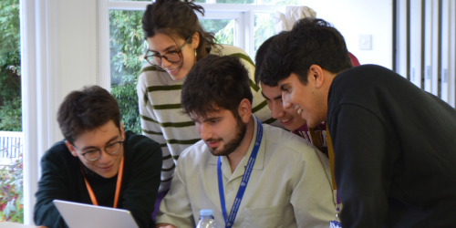 A group of five young adults gather around a laptop, engaged and smiling. They appear to be collaborating on a project, with one person seated in front of the laptop and the others leaning in, looking at the screen. The setting is the Wood-Legh room, Strathaird. There's natural light coming through large windows in the background, showing greenery outside.