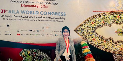 Mengxi standing in front of a large conference backdrop that reads "21st AILA World Congress: Linguistic Diversity, Equity, Inclusion, and Sustainability," held at the Kuala Lumpur Convention Centre, Malaysia, from August 11 to 16, 2024. The event celebrates 60 years of AILA (1964–2024) as part of its Diamond Jubilee. The backdrop features vibrant traditional Malaysian artwork and logos of sponsors and partners. The woman is dressed professionally in a plaid blazer and white blouse, wearing a red lanyard wi